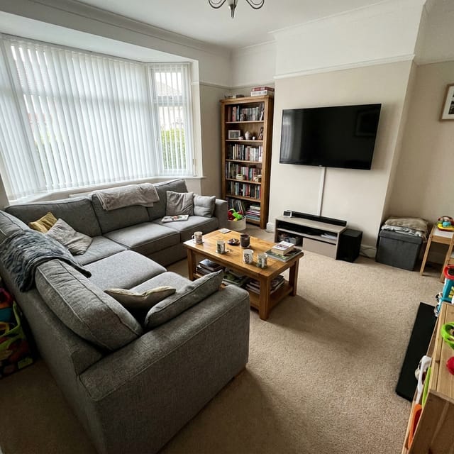 Photo of the living room: grey corner sofa, coffee table, television on the wall, bay window with blinds